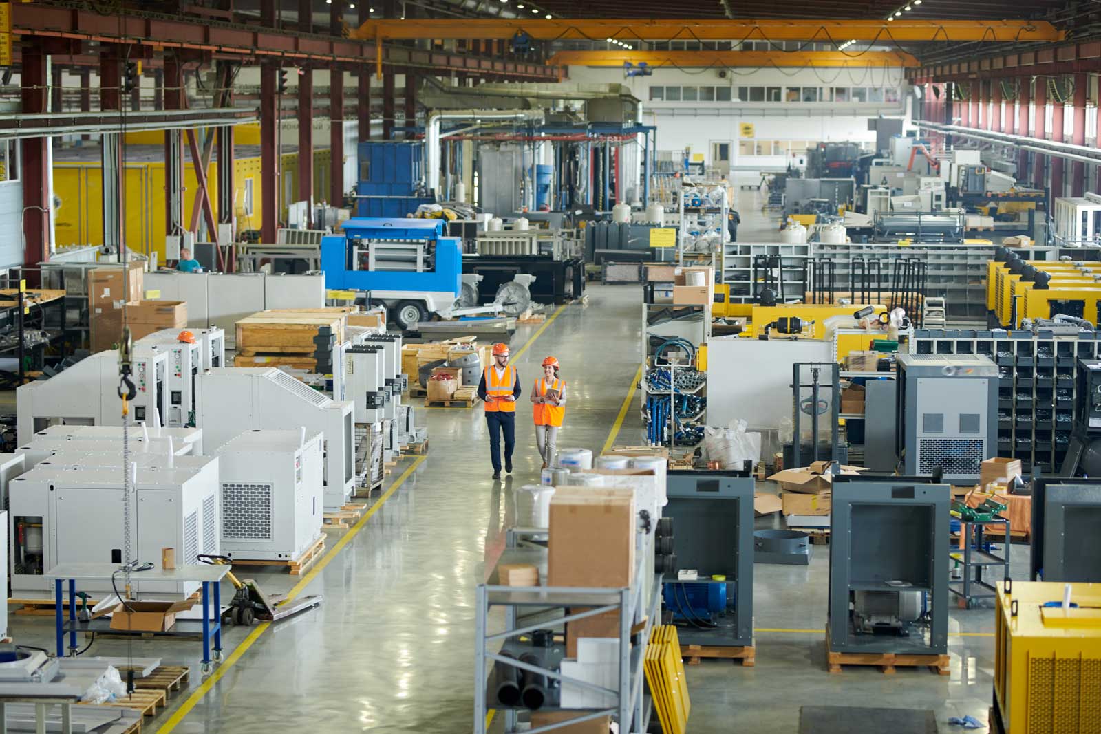 High angle full length portrait of bearded businessman wearing hardhat walking across production workshop accompanied by female factory employee, The Democratization of Manufacturing