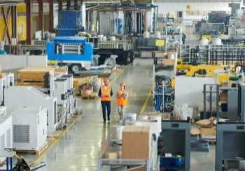 High angle full length portrait of bearded businessman wearing hardhat walking across production workshop accompanied by female factory employee, The Democratization of Manufacturing