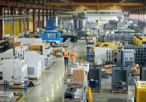 High angle full length portrait of bearded businessman wearing hardhat walking across production workshop accompanied by female factory employee, The Democratization of Manufacturing