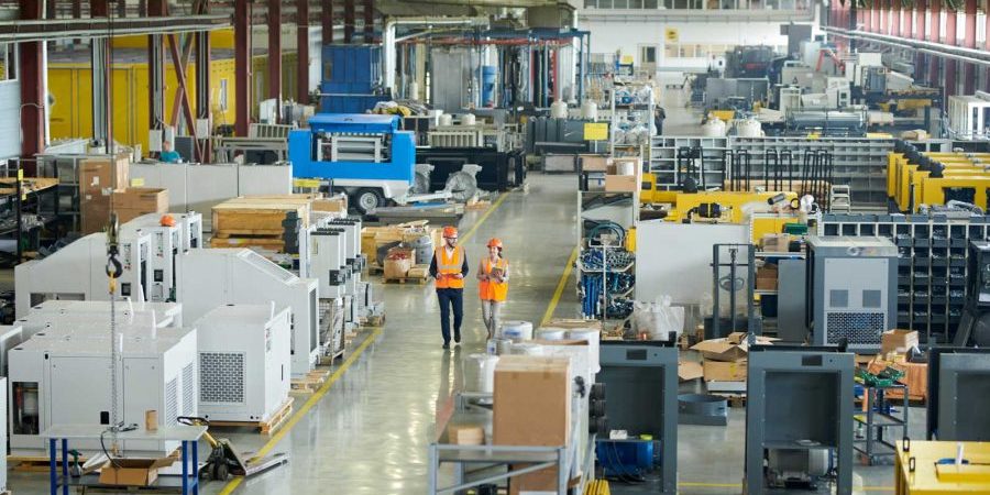 High angle full length portrait of bearded businessman wearing hardhat walking across production workshop accompanied by female factory employee, The Democratization of Manufacturing